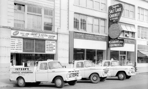 An antique image of Fetzer Architectural Woodworking cars outside of a row of buildings.